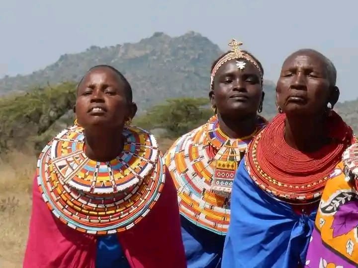 Three Maasai women in full beaded collars, standing together with the Laikipia hills behind them